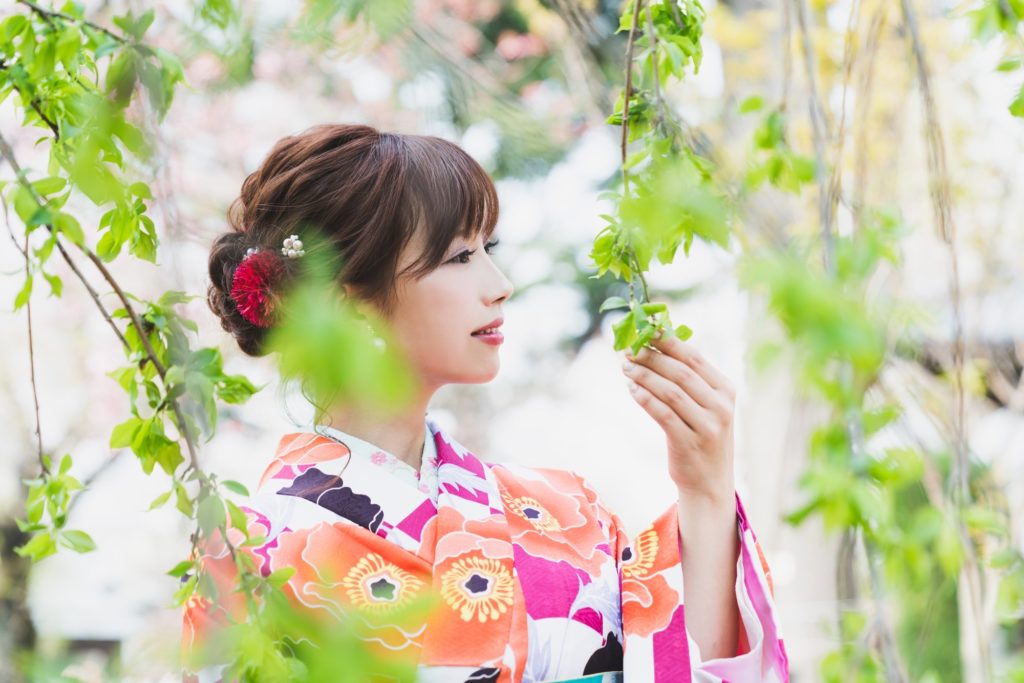 ピンクやオレンジ色の花模様の華やかな着物を着た美しい女性が若い木の葉を持って微笑んでいる、髪はやや下目に一つにまとめられ、赤い大きな花の飾りをつけている。